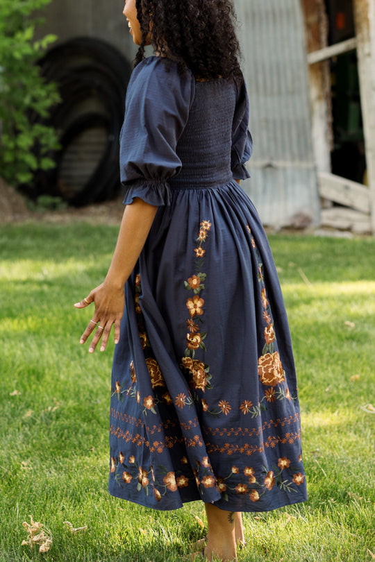 A woman with curly hair, barefoot on the grass and facing away from the camera, strolls near a corrugated metal shed and green trees. She wears the Ivy City Co Teagan Dress in Navy Embroidered Fall Floral, featuring a smocked bodice and vibrant floral patterns. The dress is maternity friendly, offering both comfort and style to her relaxed walk.