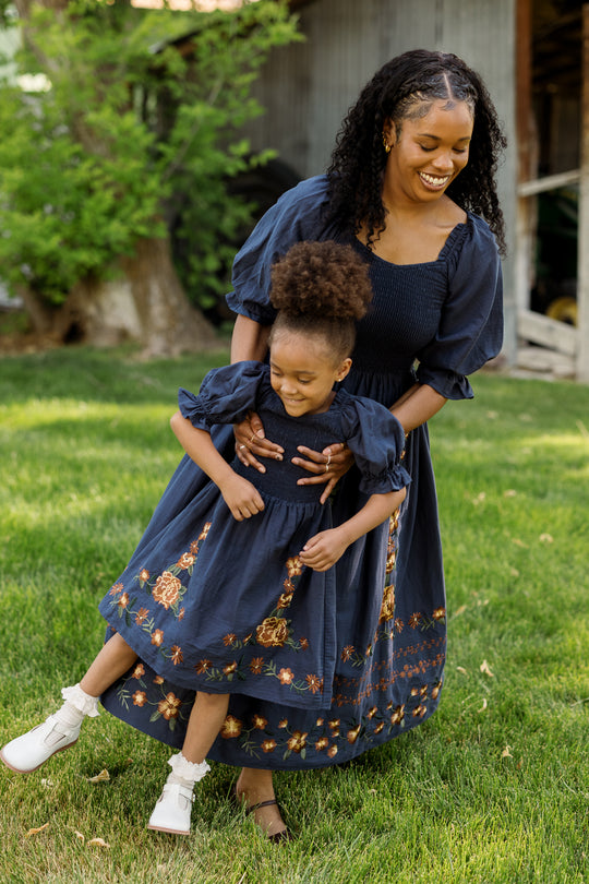 A woman and a young girl, both dressed in matching Ivy City Co Teagan Dresses in Navy Embroidered Fall Floral featuring floral embroidery and smocked bodice details, smile and play together on green grass outdoors. These charming Ivy City Co dresses are maternity friendly, making them perfect for creating special family moments.