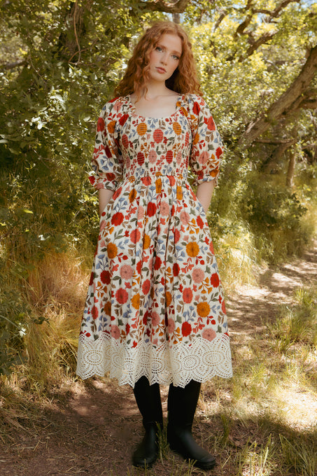 A person with long wavy red hair stands on a sunlit path, wearing Ivy City Co’s Teagan Dress in Sepia Flower—a maternity-friendly white floral midi dress with lace trim—paired with black tights, surrounded by lush green trees.