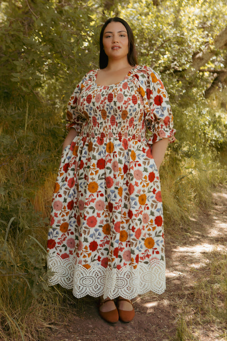 A woman stands outdoors in the Ivy City Co Teagan Dress in Sepia Flower, featuring puff sleeves and a white lace hem. Surrounded by green foliage, she shows off this floral midi style, which is also maternity friendly.
