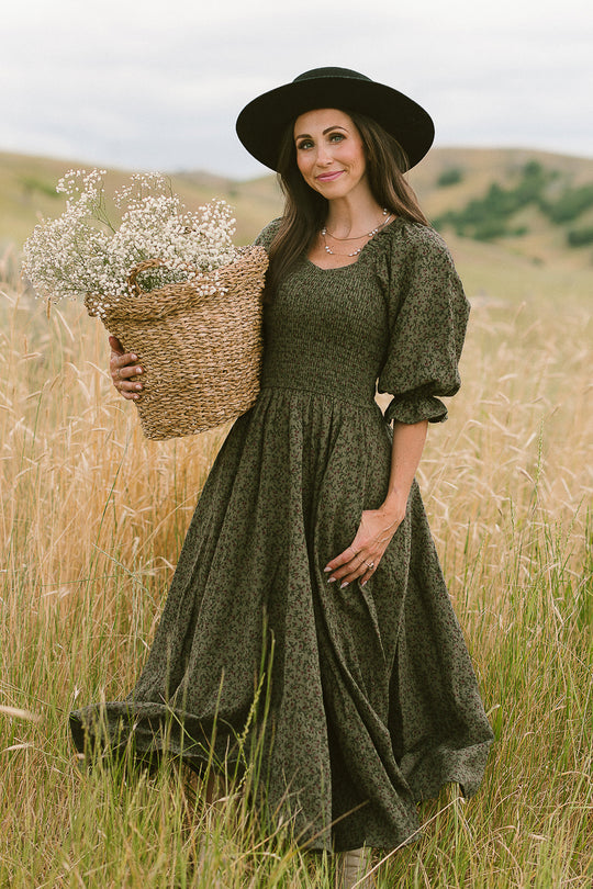 A woman wearing the Ivy City Co Teagan Dress in Woodland Floral and a black hat stands in a grassy field, holding a basket brimming with white woodland flowers. Rolling hills and an overcast sky set a magical backdrop, beautifully highlighting this maternity-friendly style.