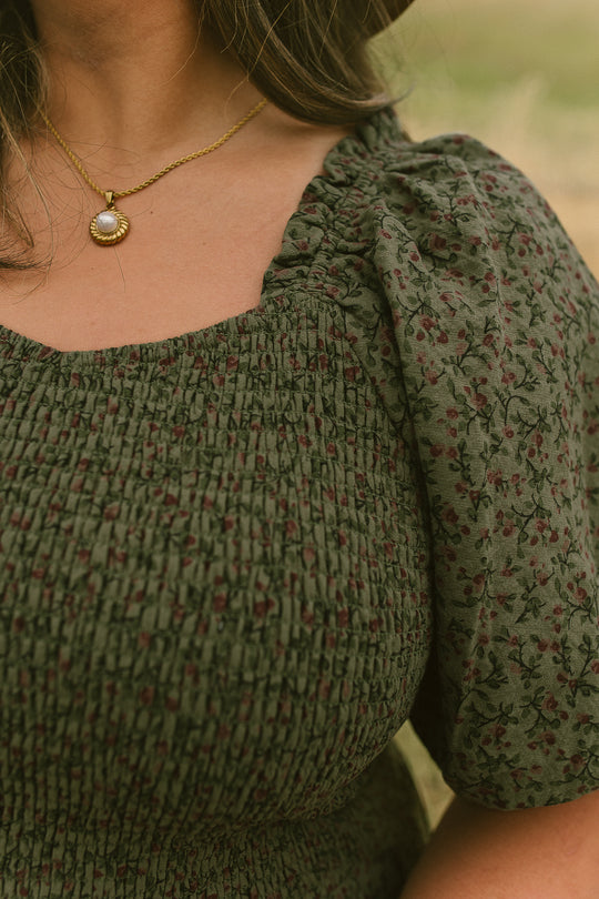 Close-up of a woman wearing the Ivy City Co Teagan Dress in Woodland Floral—a green, floral-patterned, smocked style with puff sleeves that is also maternity friendly. She accessorizes with a gold necklace featuring a round pendant, while her face is partially out of frame.