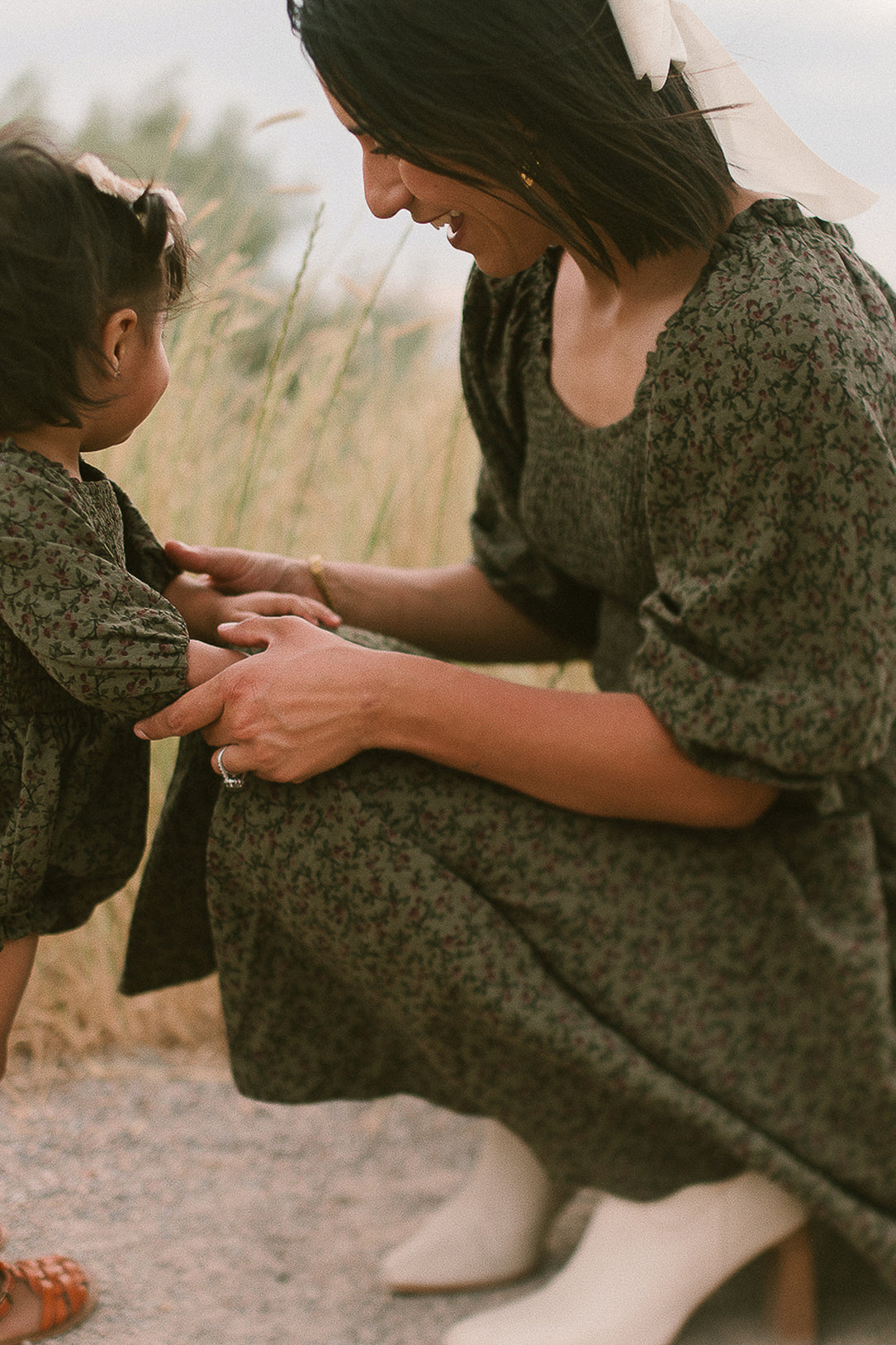 A woman kneels and smiles at a toddler as they hold hands outdoors, both dressed in matching Ivy City Co Teagan Dresses in Woodland Floral. Perfect for a maternity-friendly look, the woman’s hair is adorned with a white ribbon, and grass is visible in the background.