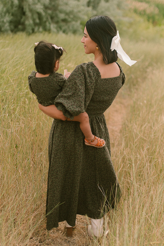 A woman with a white ribbon in her hair carries a small child through a grassy field. Both are wearing matching Ivy City Co Teagan Dresses in the Woodland Floral pattern. Their outfits are stylish and maternity-friendly as they walk away on a narrow path surrounded by tall grass.
