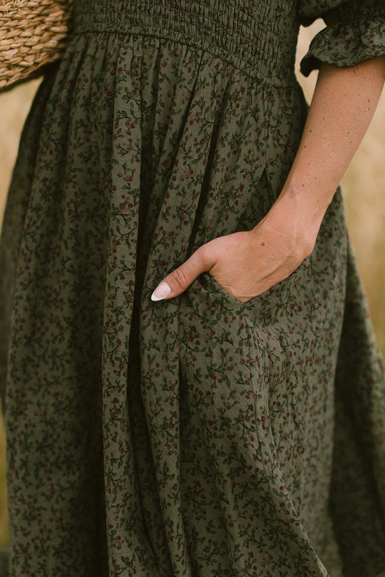 A person wearing Ivy City Co's Teagan Dress in Woodland Floral, featuring a maternity-friendly gathered waist, stands with one hand in their pocket. Only the lower torso and hand are visible against a blurred background.