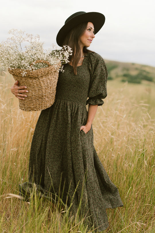 A woman wearing the Ivy City Co Teagan Dress in Woodland Floral and a black hat stands in a grassy field, holding a large wicker basket filled with white flowers. The maternity friendly dress flows elegantly as hills and a cloudy sky appear in the background.