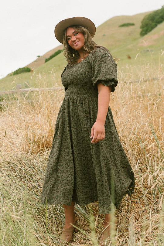 A woman in a long, dark Teagan Dress in Woodland Floral by Ivy City Co and a tan hat stands in a field of tall grass, smiling at the camera. This maternity-friendly style from Ivy City Co pairs beautifully with the rolling green hills visible in the background.