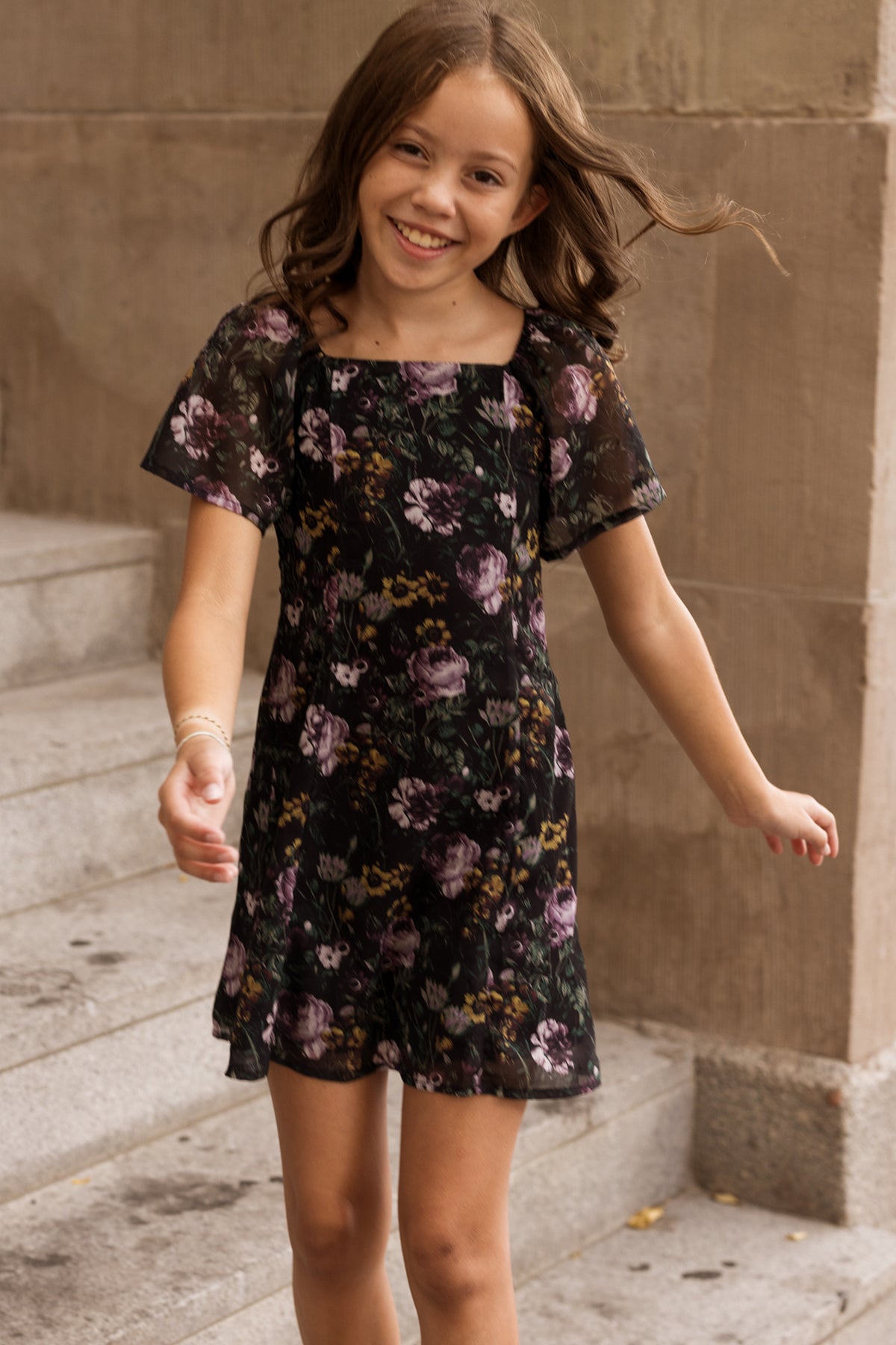 A smiling girl with wavy brown hair wears the Ivy City Co Teen Blakely Dress in Belladonna Bloom as she walks outside near stone steps and a beige wall.