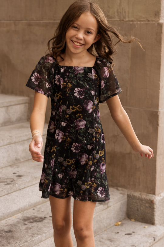 A smiling girl with wavy brown hair wears the Ivy City Co Teen Blakely Dress in Belladonna Bloom as she walks outside near stone steps and a beige wall.