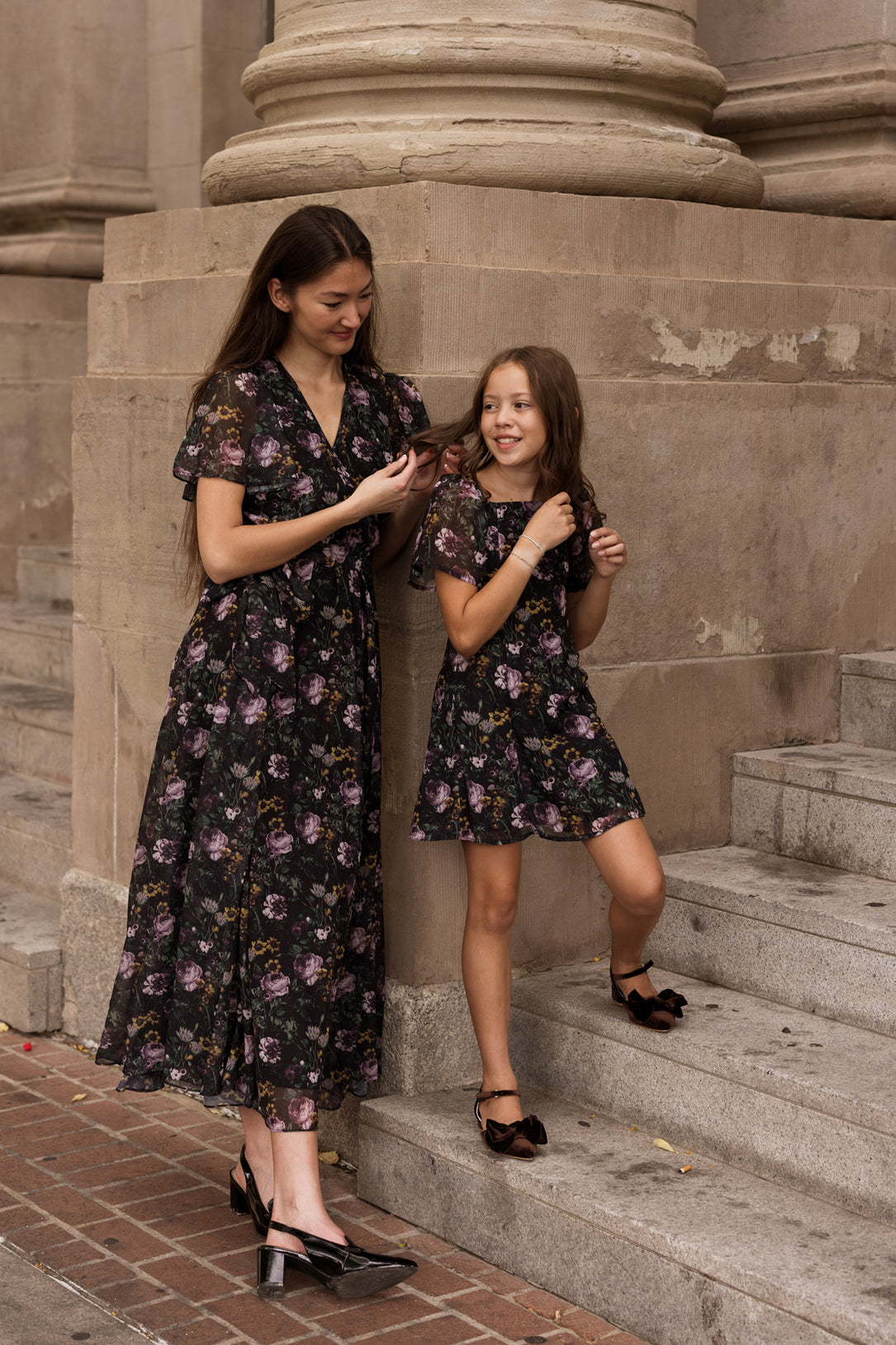 A woman and a girl stand on stone steps outdoors wearing matching floral dresses from Ivy City Co. The woman wears a flutter sleeve dress, looking at her phone, while the girl beams in the Teen Blakely Dress in Belladonna Bloom, one foot playfully perched on a step.
