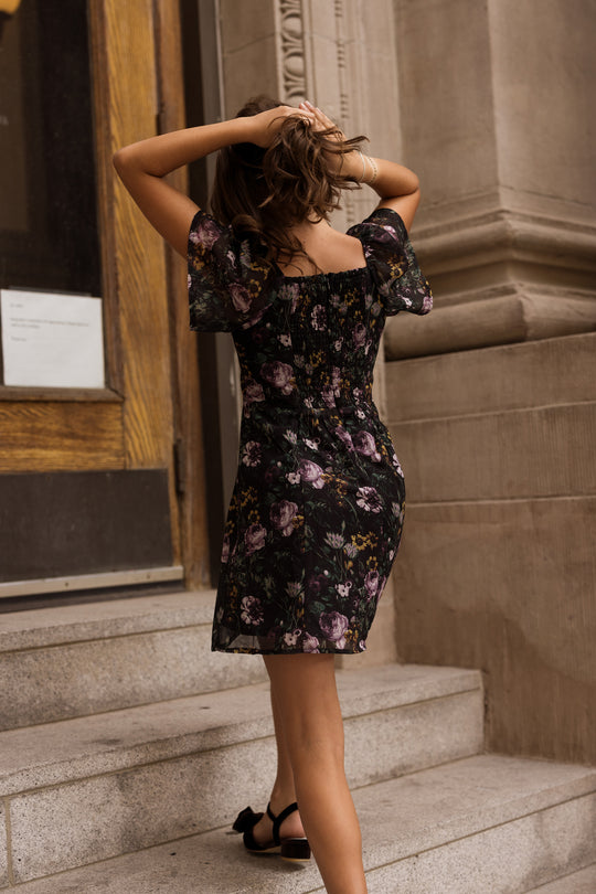 A woman in the Ivy City Co Teen Blakely Dress in Belladonna Bloom and black sandals stands on stone steps, facing away as she adjusts her hair, near a wooden door and stone building.