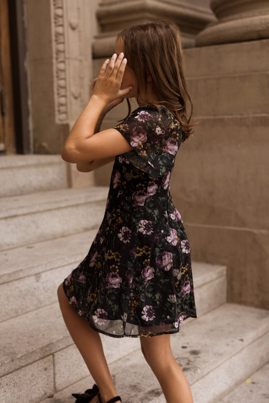A girl in the Ivy City Co Teen Blakely Dress in Belladonna Bloom walks up stone steps, covering her face with both hands.