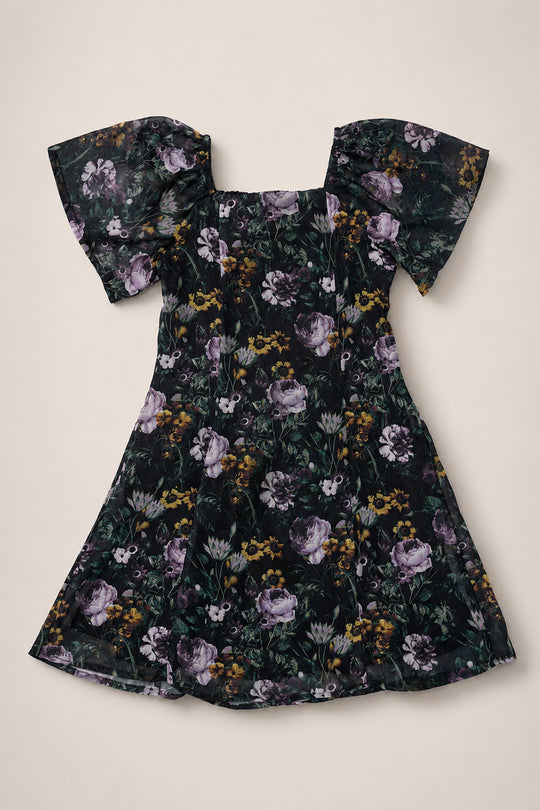 The Ivy City Co Teen Blakely Dress in Belladonna Bloom, a black girls’ knee-length dress with a floral print in purple, green, and yellow, featuring puffed short sleeves and a square neckline, is laid flat on a cream background.
