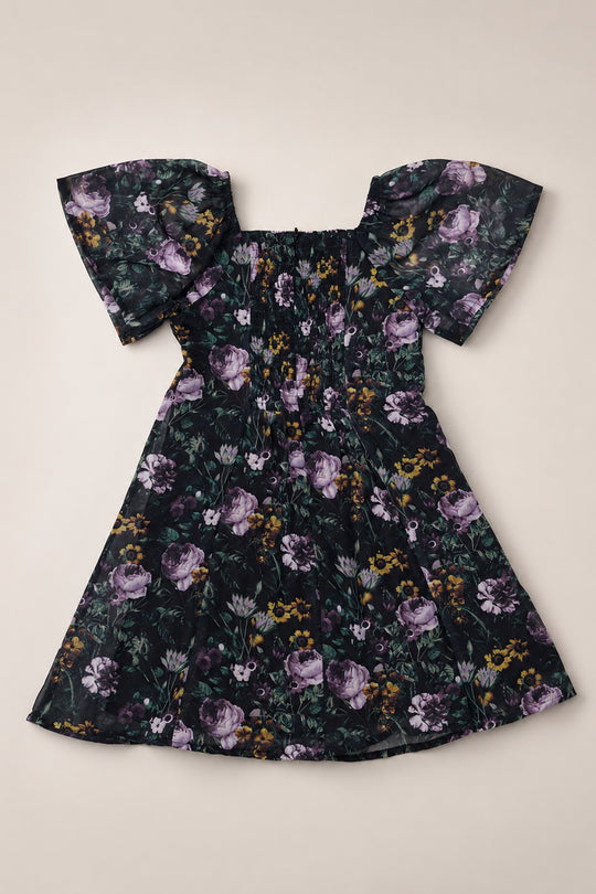 The Ivy City Co Teen Blakely Dress in Belladonna Bloom showcases a black background with a striking purple floral print, short off-shoulder flutter sleeves, a fitted ruched bodice, and a flared knee-length skirt—an ideal choice for those seeking a stylish girls’ dress.