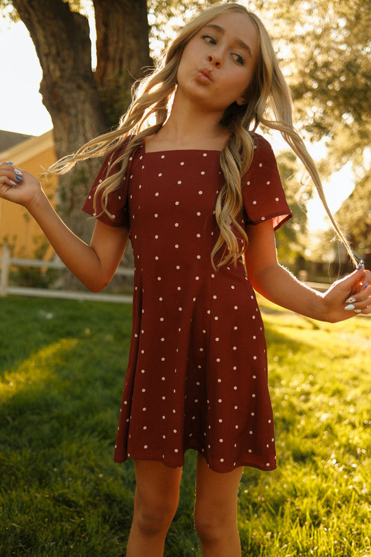 A young girl with long blonde hair stands outdoors in the Ivy City Co Teen Blakely Dress in Cranberry Dainty Dot, playfully holding her hair and making a pouty face. Charming petal sleeves add detail to the dress, while sunlight streams through the trees behind her.