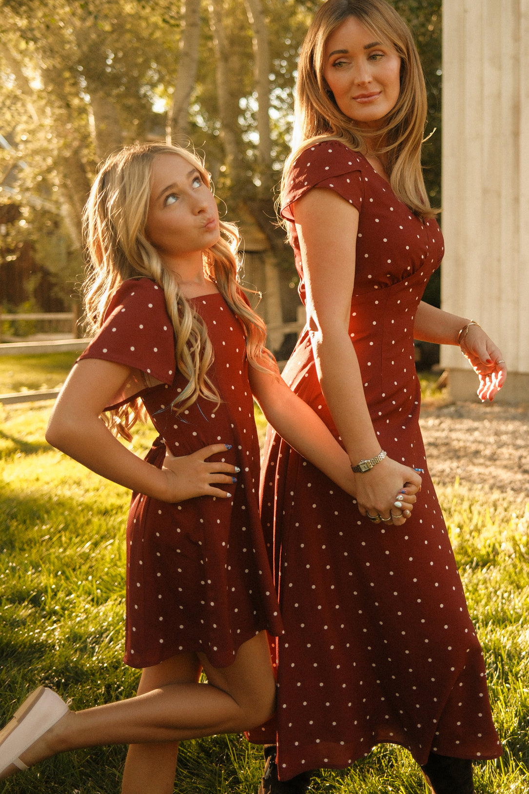 A woman and a girl wearing matching maroon polka dot dresses hold hands outdoors. The girl is dressed in the Ivy City Co Teen Blakely Dress in Cranberry Dainty Dot, featuring playful petal sleeves and a graceful midi-length hem, as she poses playfully while the woman stands beside her, both bathed in warm sunlight.