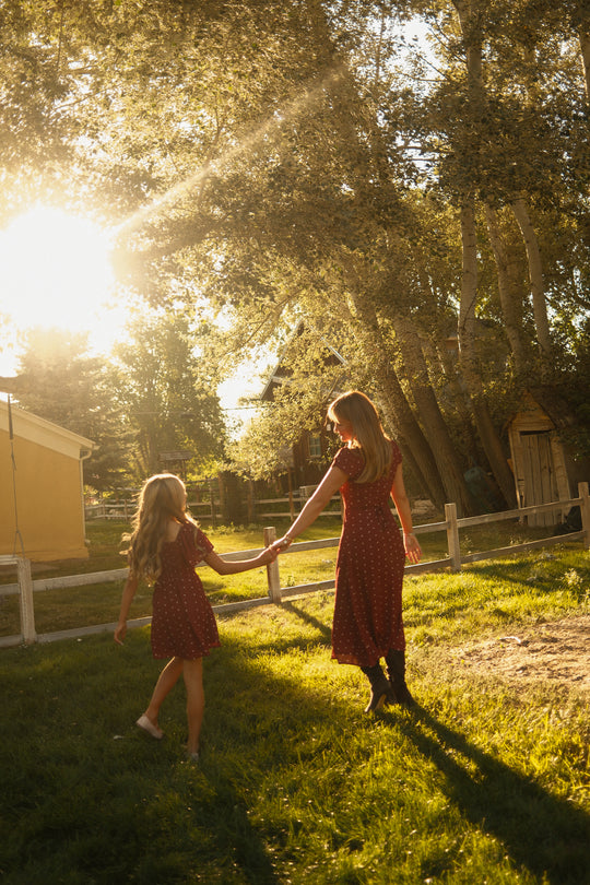 Two girls wear matching Ivy City Co Teen Blakely Dresses in Cranberry Dainty Dot, featuring petal sleeves and midi-length skirts, as they hold hands and walk across sunlit grass surrounded by tall trees and wooden fences, bathed in warm golden light.
