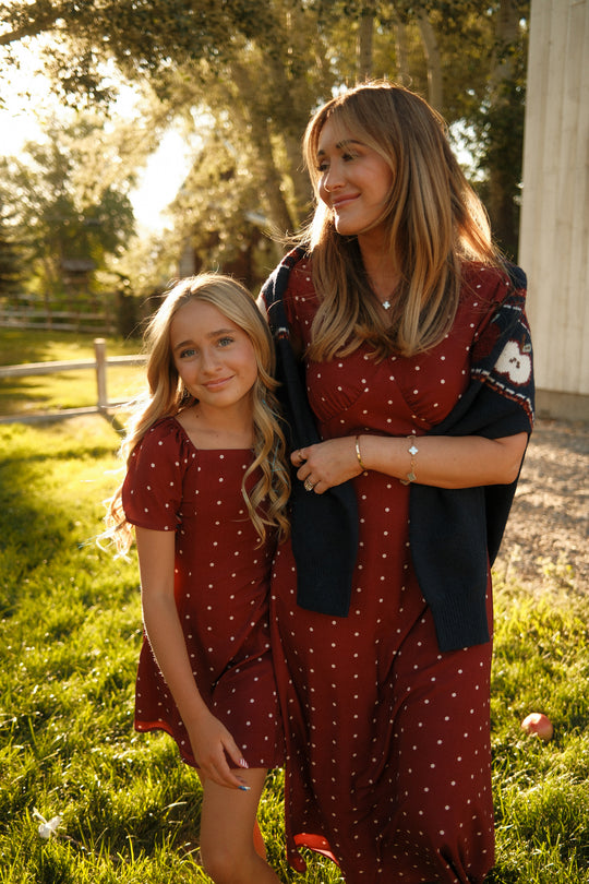 Two people wearing matching Ivy City Co Teen Blakely Dresses in Cranberry Dainty Dot with charming petal sleeves stand close together outside on a sunny day, smiling and posing affectionately with trees and sunlight in the background.