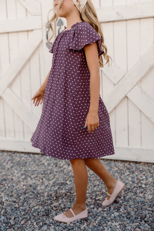 A girl wearing the Ivy City Co Teen Blakely Dress in Aubergine Ditsy Dot, featuring a smocked bodice and flutter sleeves, paired with pink shoes, walks on gravel in front of a white wooden gate, facing sideways with her head slightly turned.