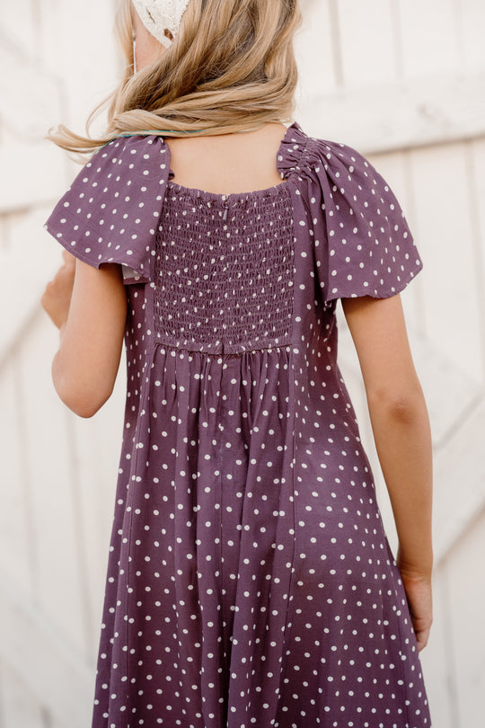 A young girl with light hair stands with her back to the camera in front of a light wooden fence, wearing the Ivy City Co Teen Blakely Dress in Aubergine Ditsy Dot, which features a smocked bodice, delicate cap sleeves, and a charming purple-and-white polka dot pattern.