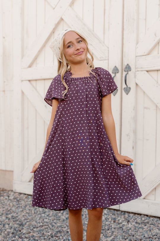 A smiling young girl stands on gravel in front of a white wooden gate, holding out her Ivy City Co Teen Blakely Dress in Aubergine Ditsy Dot, which features a smocked bodice and flutter sleeves.