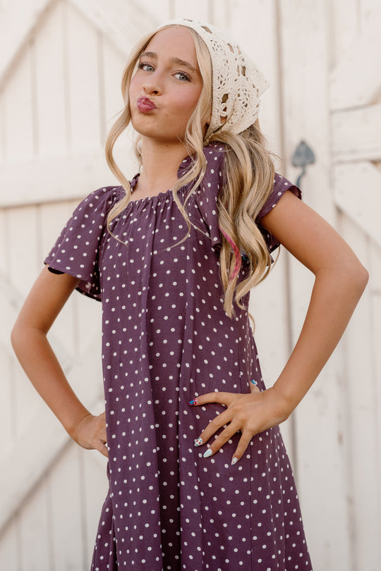 A young girl with long blond hair stands with her hands on her hips and puckered lips in front of a light wooden background. She is wearing the Ivy City Co Teen Blakely Dress in Aubergine Ditsy Dot, which features flutter sleeves and a smocked bodice. Her charming look is completed with a white crocheted headscarf.