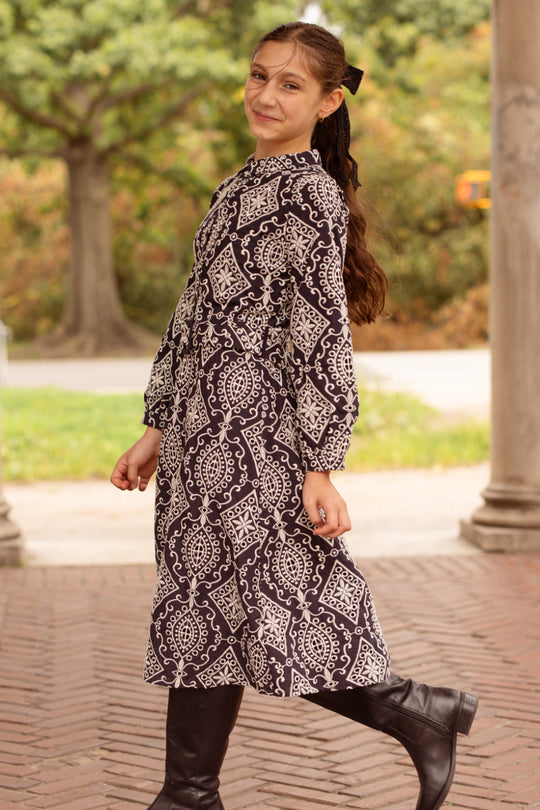A young girl smiles outdoors, wearing the Ivy City Co Teen Clover Dress in Midnight Tile—a long-sleeve black and white paisley midi dress with a belt—paired with black knee-high boots, her dark hair pulled back in a ponytail adorned with a black bow.