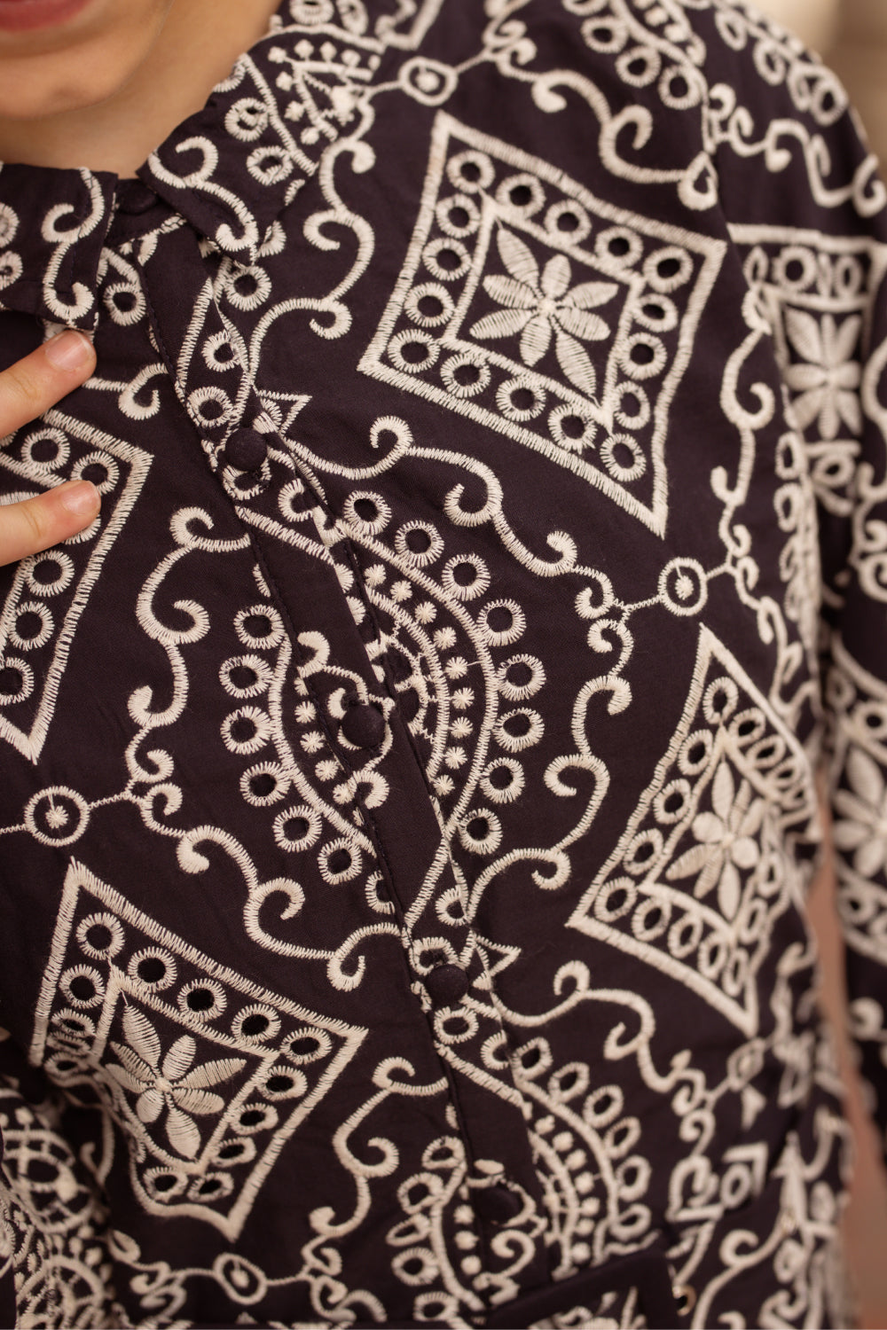 Close-up of a person wearing the Ivy City Co Teen Clover Dress in Midnight Tile, featuring intricate white embroidery in geometric and floral patterns; their hand is near the collar.