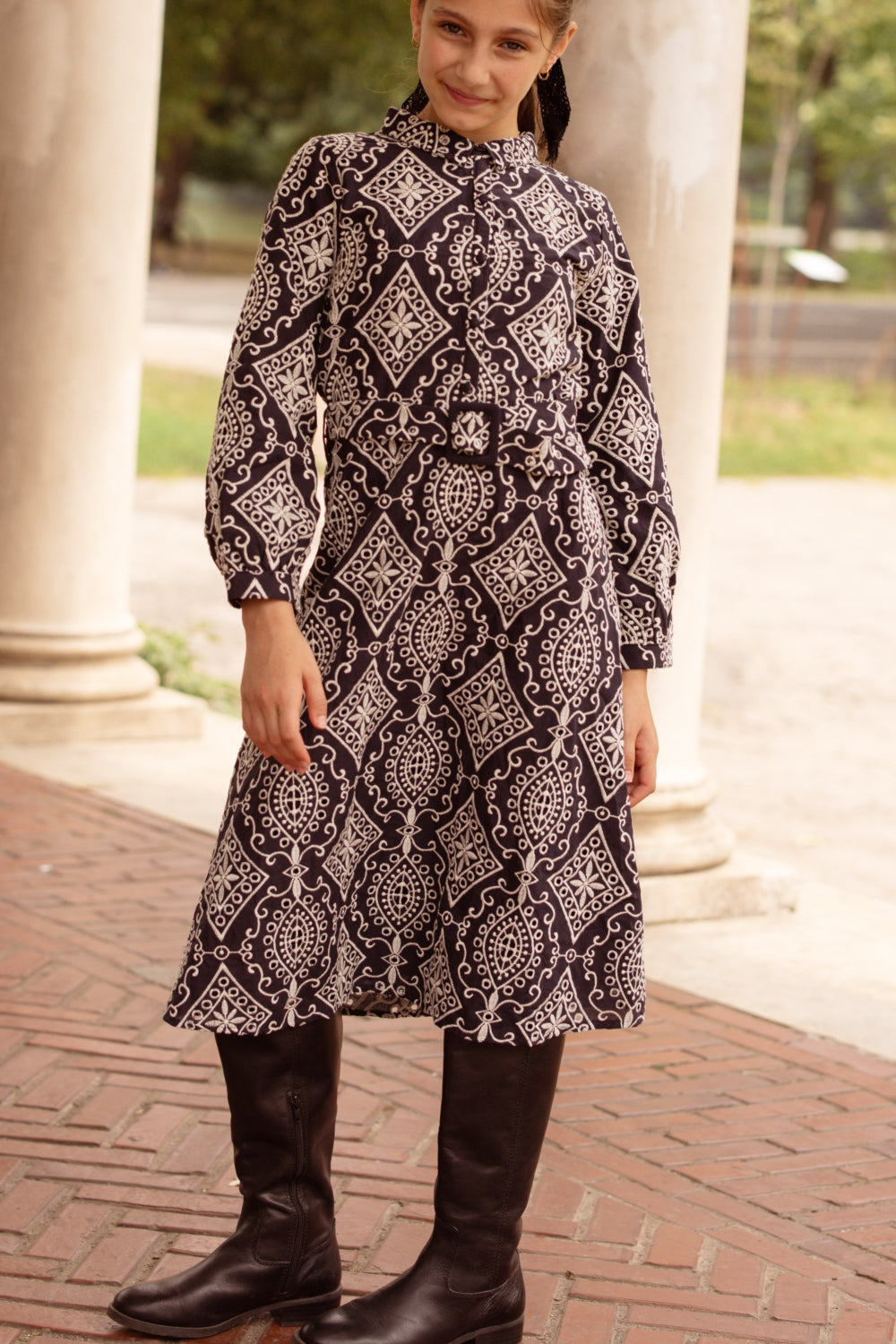 A young girl stands outside on a brick walkway, wearing the Ivy City Co Teen Clover Dress in Midnight Tile and tall dark boots, smiling slightly at the camera with one hand on her hip and columns in the background.