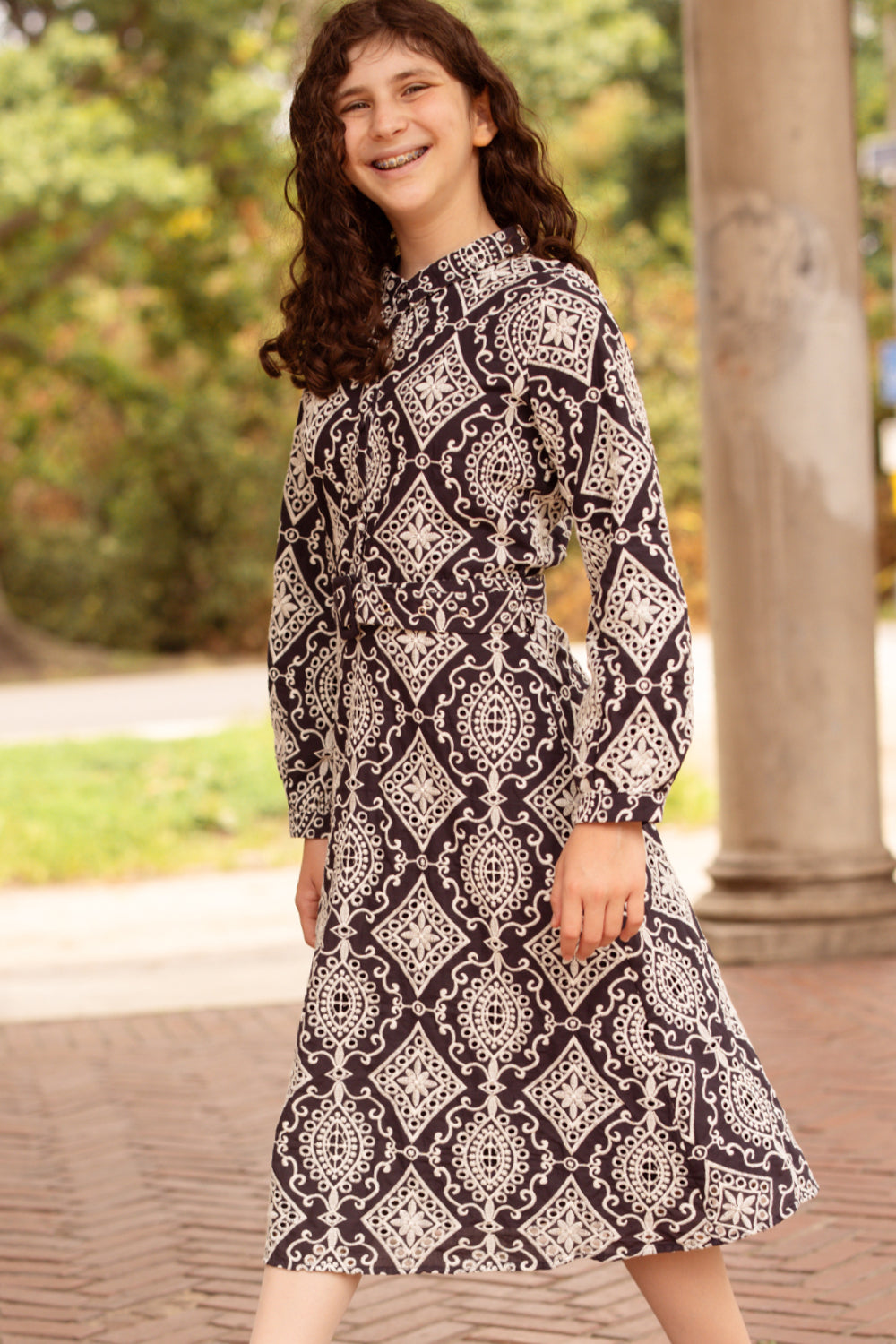 A person with long curly hair, wearing the Ivy City Co Teen Clover Dress in Midnight Tile, stands outdoors on a brick path, smiling. Green trees and a column are visible in the background.