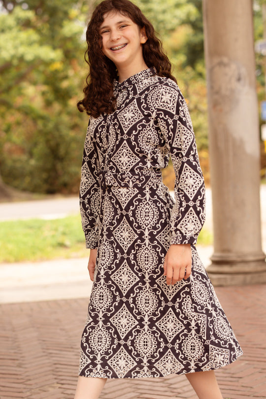 A person with long curly hair, wearing the Ivy City Co Teen Clover Dress in Midnight Tile, stands outdoors on a brick path, smiling. Green trees and a column are visible in the background.