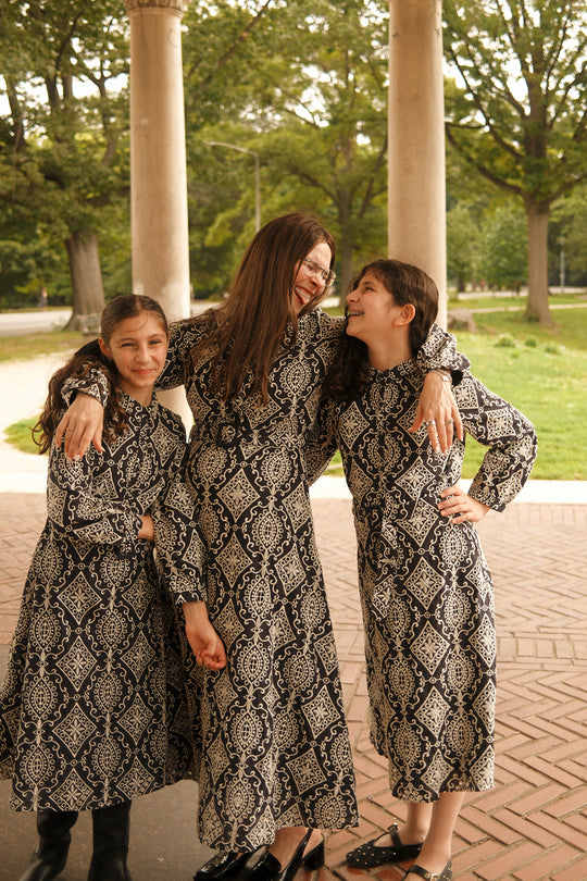 Three people wearing matching black and white patterned dresses—including the Ivy City Co x Miriam Midnight Tile midi-length dress and the Ivy City Co Teen Clover Dress in Midnight Tile—stand under a pavilion, smiling and laughing together in a park surrounded by green trees.