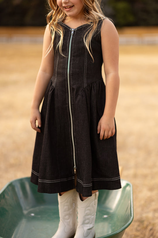 A young girl stands outdoors in a green wheelbarrow on a field, wearing the Ivy City Co Teen Edith Dress in Black—a sleeveless black midi dress with a front zipper—and white cowboy boots.