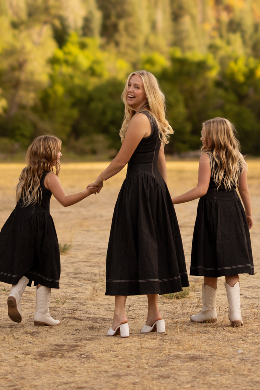 Three people—a woman and two young girls—all wearing the Ivy City Co Teen Edith Dress in Black, paired with white boots, hold hands as they walk together outdoors on dry grass with trees in the background.