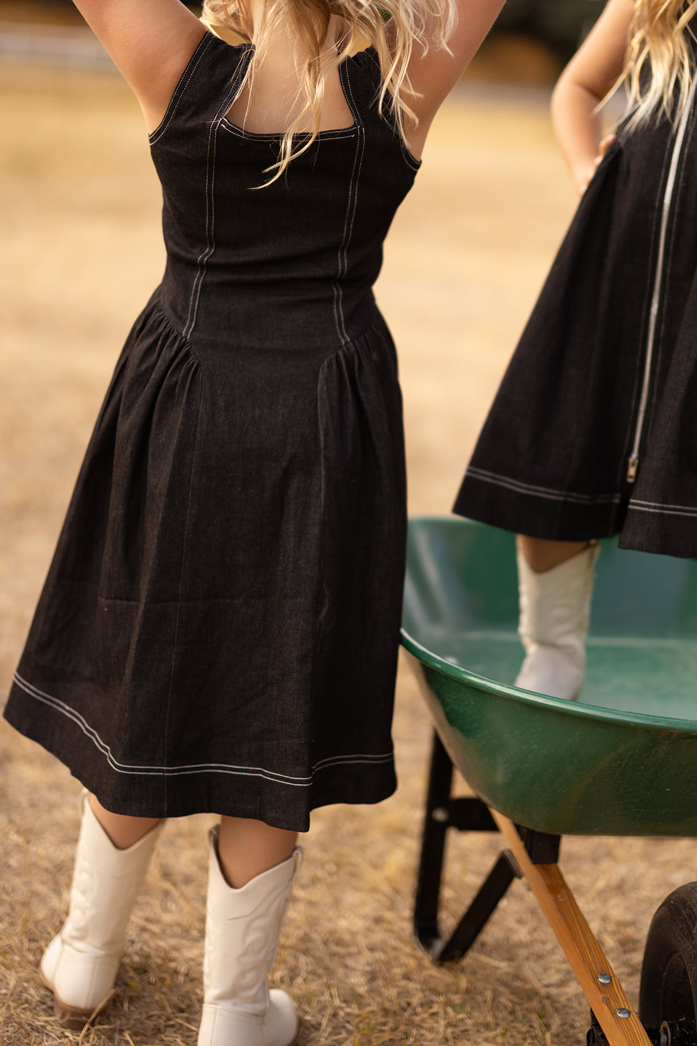 A young child in the Ivy City Co Teen Edith Dress in Black and white cowboy boots stands beside a green wheelbarrow outdoors on dry grass, raising their arms.