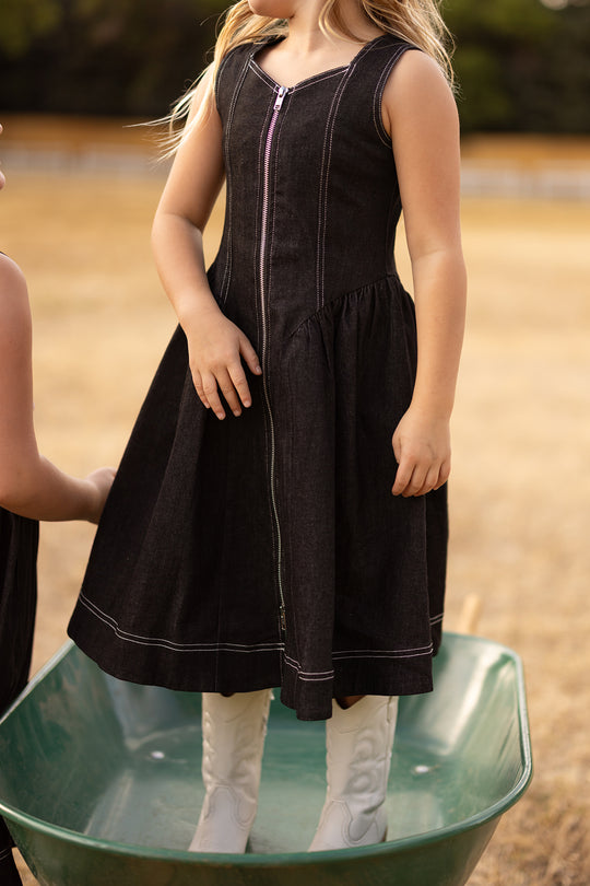 Wearing the Ivy City Co Teen Edith Dress in Black, a girl stands in a green wheelbarrow on dry grass, paired with white cowboy boots; another child’s arm is visible beside her.