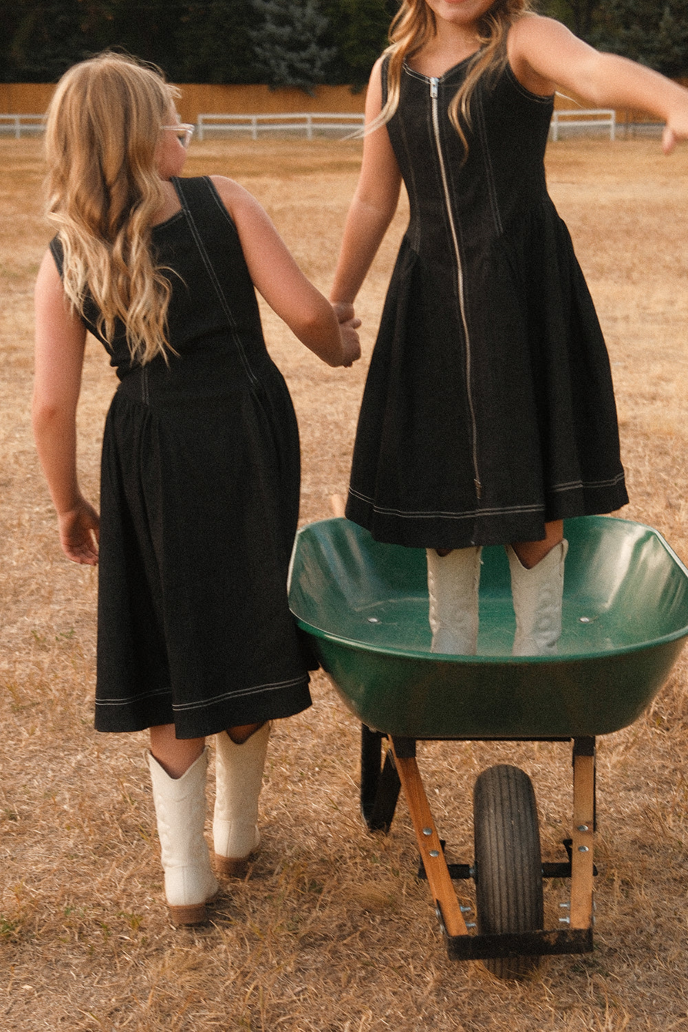Two girls in Ivy City Co’s Teen Edith Dress in Black and white boots stand in a dry, grassy field. One balances playfully inside a green wheelbarrow while the other supports her with an outstretched hand. With their long, wavy blonde hair and chic cotton-spandex dresses, the scene is filled with effortless style and youthful charm.
