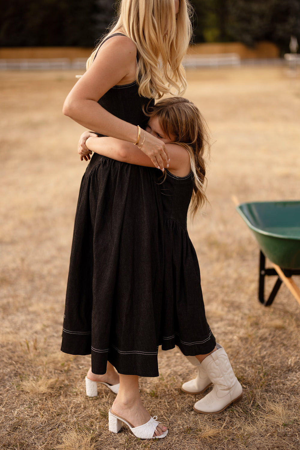 A young girl in white boots hugs an adult woman around the waist outdoors. Both are wearing chic black midi dresses, with the girl dressed in Ivy City Co’s charming Teen Edith Dress in Black. The scene unfolds in a dry, grassy field, with a green wheelbarrow partially visible nearby.