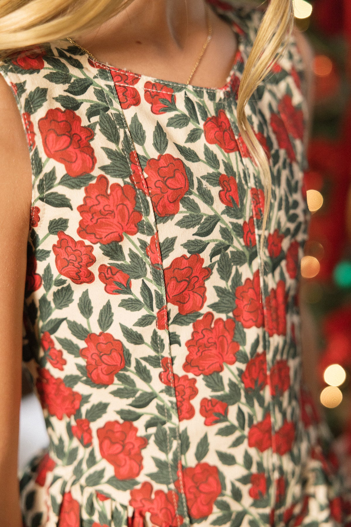 A close-up of the Ivy City Co Teen Edith Dress in Red Roses, featuring a vibrant red rose and green leaf pattern, worn by a person with long blond hair. Warm, festive lights blur in the background, making this dress a perfect choice for holiday parties.