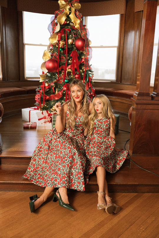 Two people, both with long blonde hair, are smiling and posing together in a cozy, wood-paneled room decorated for Christmas. They are wearing matching Ivy City Co Teen Edith Dresses in Red Roses as they sit in front of a Christmas tree with gifts underneath.