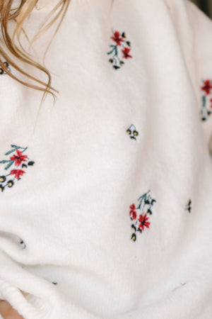 A close-up of the Ivy City Co Teen Ellis Sweater in Ivory Jubilant Bloom shows its delicate, holiday-inspired red and green embroidery. The sweater’s soft white knit is paired with a hint of a girl’s wavy hair and neck at the top left, highlighting the cozy charm of this festive piece.