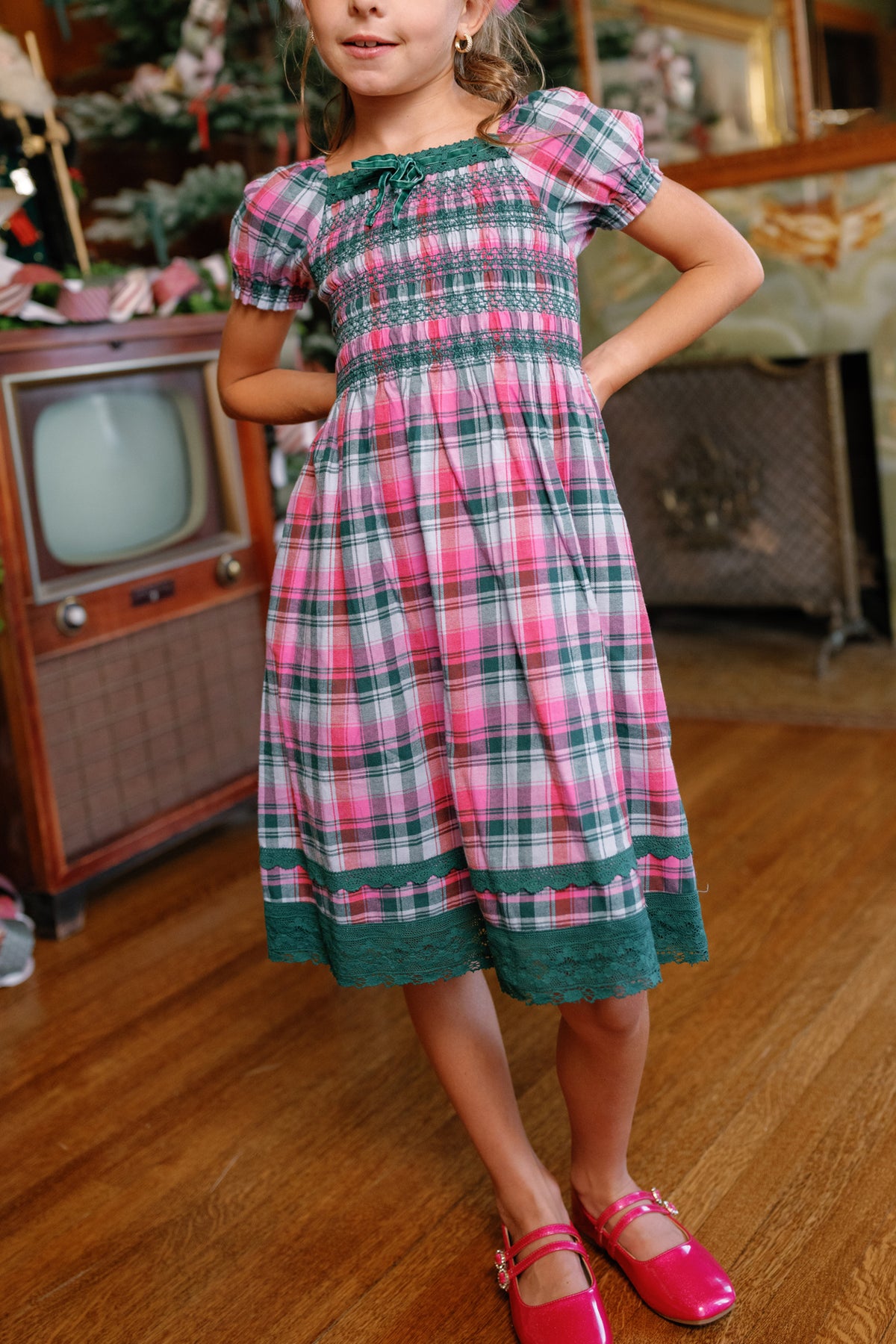 A young girl indoors stands with her hands on her hips, wearing the Ivy City Co Teen Sunday Best Dress in Vibrant Plaid and shiny pink shoes. In the background are an old-fashioned TV and a decorated Christmas tree.