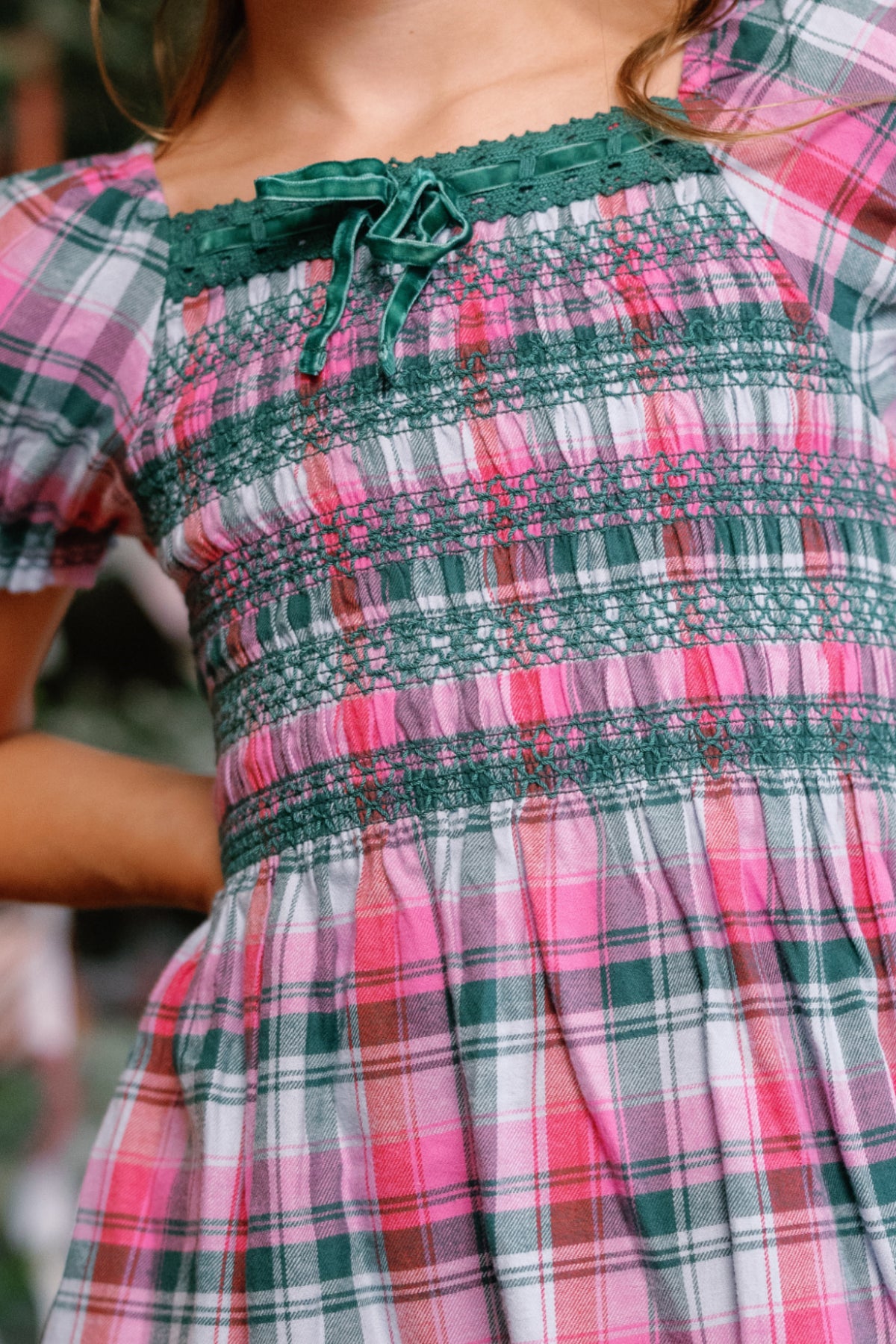 A child wearing the Ivy City Co Teen Sunday Best Dress in Vibrant Plaid, featuring green smocking and a small green bow at the neckline—a festive choice for Christmas.