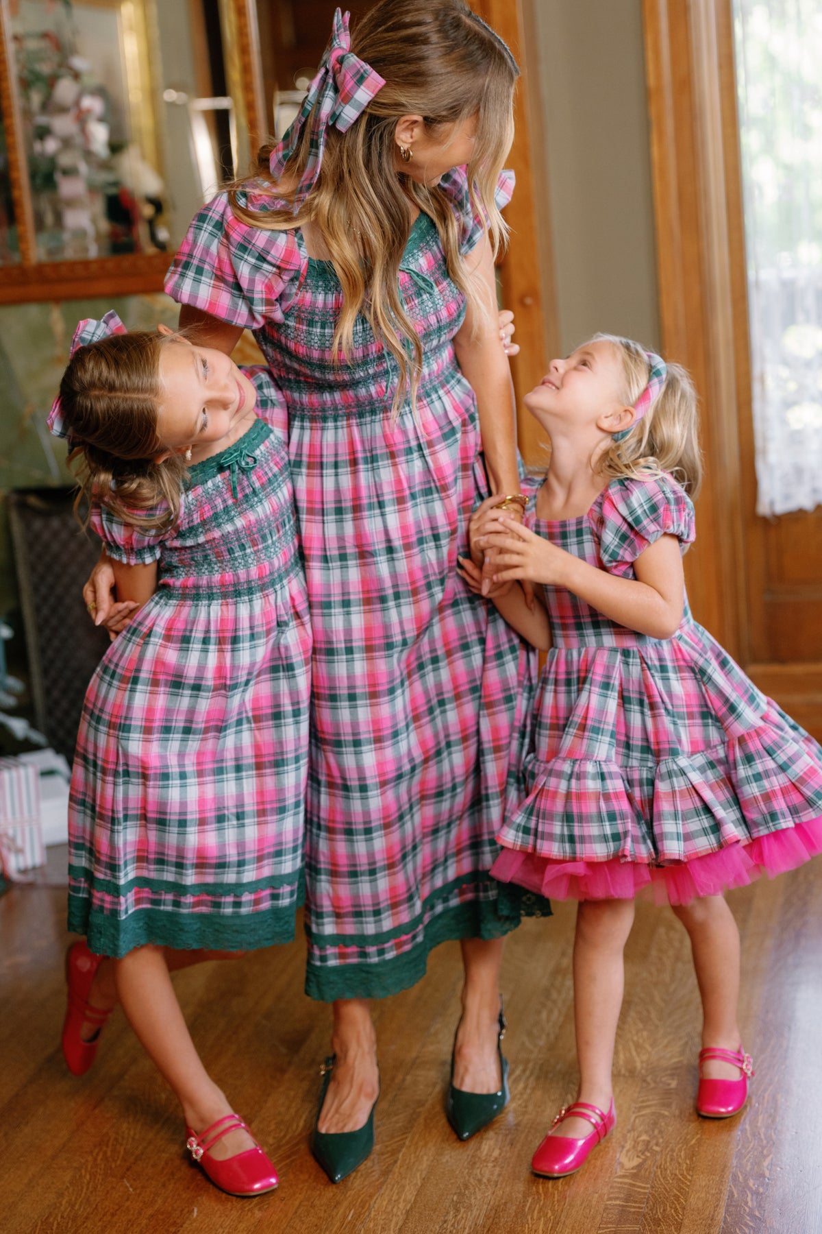 A woman and two young girls, all dressed in Ivy City Co’s matching Teen Sunday Best Dress in Vibrant Plaid, stand smiling together indoors on a wooden floor. The girls wear pink shoes and hold the woman’s hands, their cheerful plaid outfits making the perfect festive look for family celebrations this Christmas.