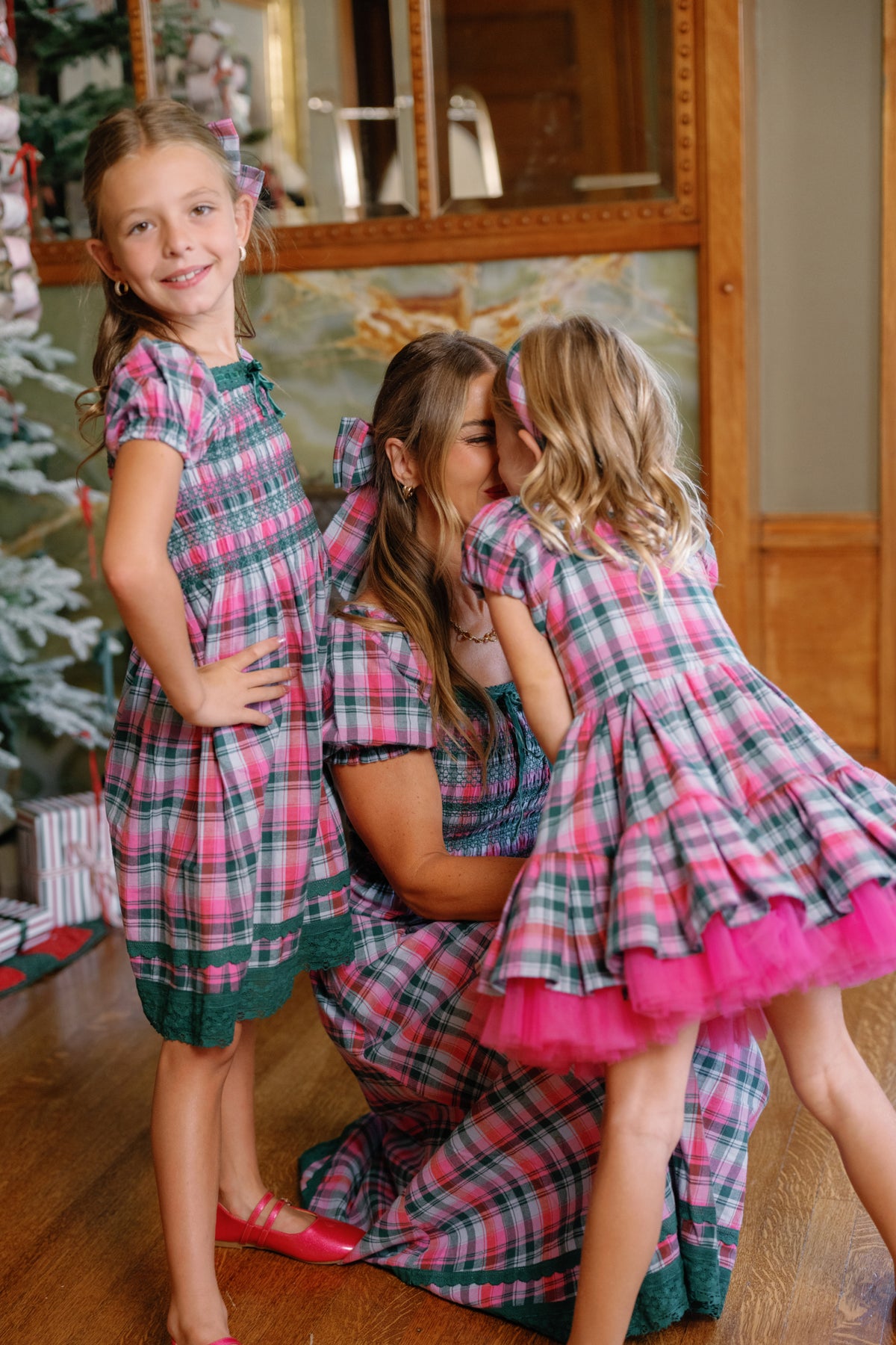 A woman kneels beside a Christmas tree with wrapped presents as one girl kisses her cheek and another stands smiling; all three wear matching Ivy City Co Teen Sunday Best Dresses in Vibrant Plaid.
