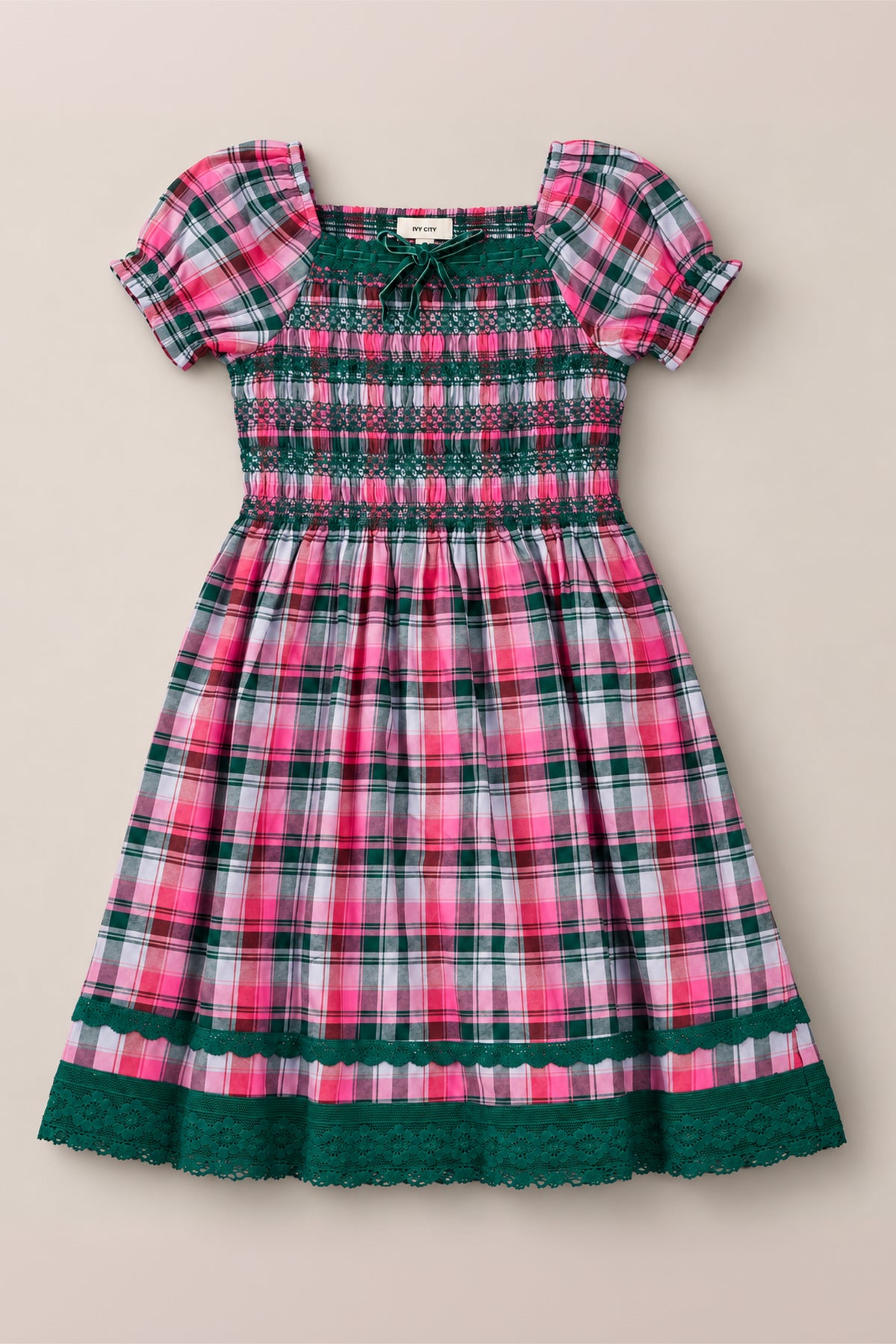 The Ivy City Co Teen Sunday Best Dress in Vibrant Plaid is a short-sleeve, knee-length dress featuring a pink, green, and white plaid pattern with a smocked cotton bodice, green lace trim, and ruffle accents at the hem and sleeves—perfect for festive Christmas celebrations or any cheerful occasion.