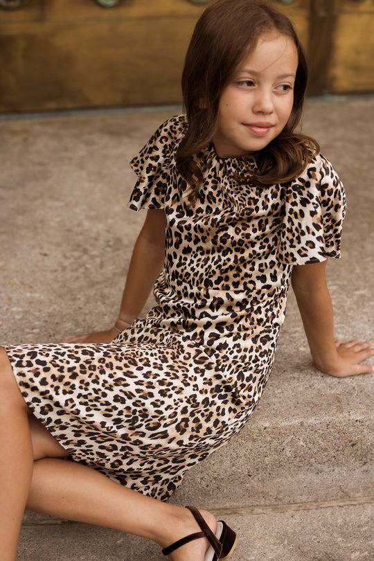 A young girl with brown hair sits on concrete steps, wearing the Ivy City Co Teen Tallulah Dress in Leopard and black sandals, looking to the side with a slight smile.