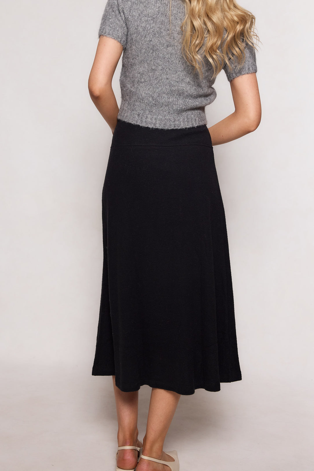 A woman with long blonde hair stands with her back to the camera, wearing a short-sleeve gray sweater, beige sandals, and the Tenley Skirt in Black by Ivy City Co.