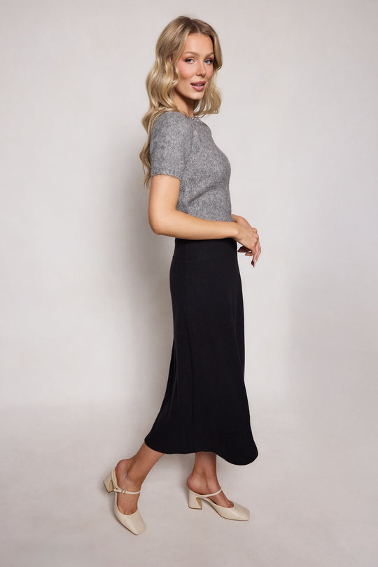 A woman with long blonde hair stands in profile against a light background, smiling softly. She wears a grey short-sleeve sweater, cream low-heeled shoes, and the Ivy City Co Tenley Skirt in Black.