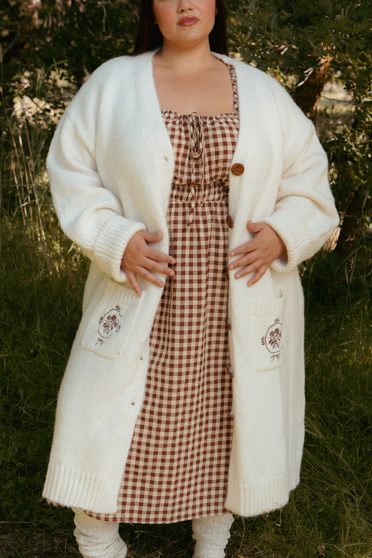 A woman stands outdoors in the Ivy City Co Teresa Cardigan in Cream, a long, nursing-friendly maternity cardigan with floral embroidery on the pockets, layered over a brown and white checkered dress as she gently rests her hands on her stomach.
